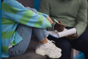 picture of a teenager sitting with their knees clutched against their chest and talking with an adult male who has a notepad and is writing notes