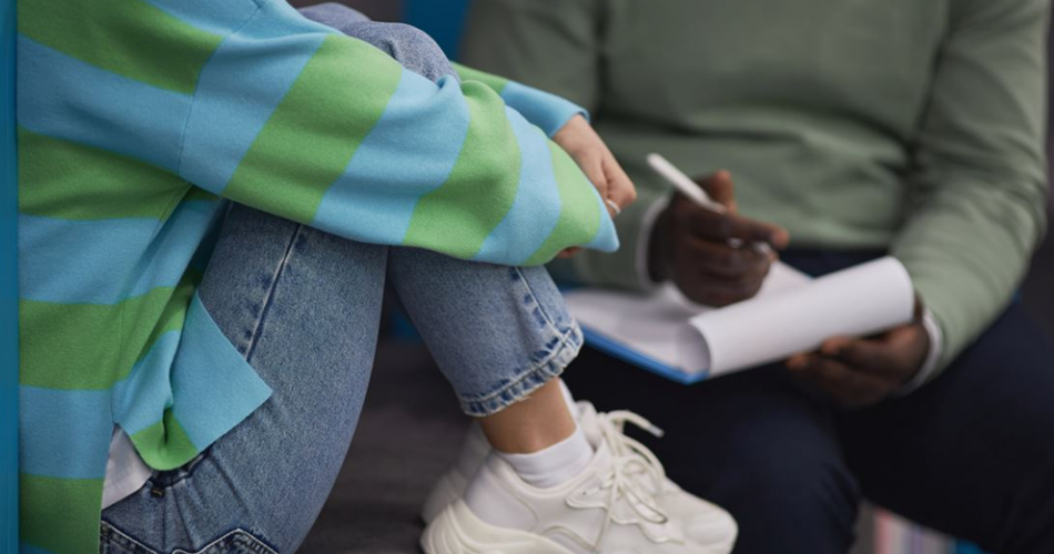 picture of a teenager sitting with their knees clutched against their chest and talking with an adult male who has a notepad and is writing notes