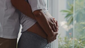 Pregnant woman standing in front of window with her partner hugging her stomach from behind.