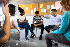 Teen students sit in chairs in a semi-circle with one student offering another a supportive hand.