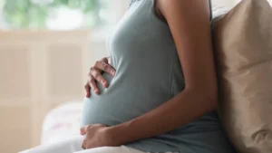 Pregnant woman sitting on a couch holding her stomach in both hands with a window in the background.