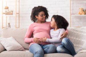 Pregnant woman sitting on a couch smiling down at her daughter sitting next to her.