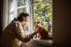 Young adult petting a cat sitting on a windowsill.