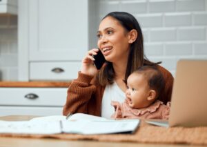 Mother sits at a table holding her baby while talking on a phone.