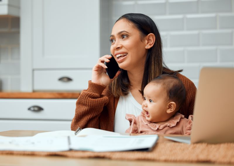 Mother sits at a table holding her baby while talking on a phone.