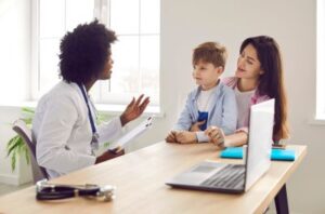 A doctor sits at a desk across from a mother with her son on her lap as they have a discussion.