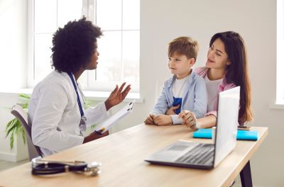A doctor sits at a desk across from a mother with her son on her lap as they have a discussion.