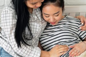 A pregnant woman sitting next to her mother as they both smile and hold her stomach.