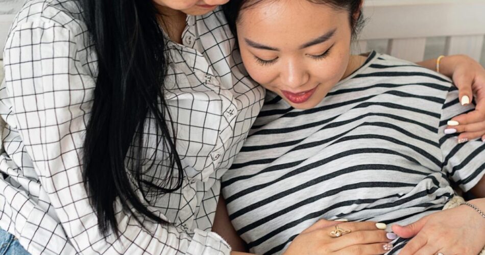 A pregnant woman sitting next to her mother as they both smile and hold her stomach.