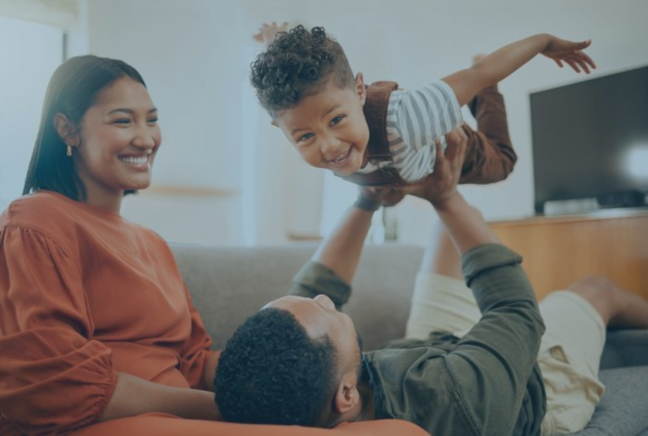 A mom sits on a couch smiling at her small child being held up by the father.