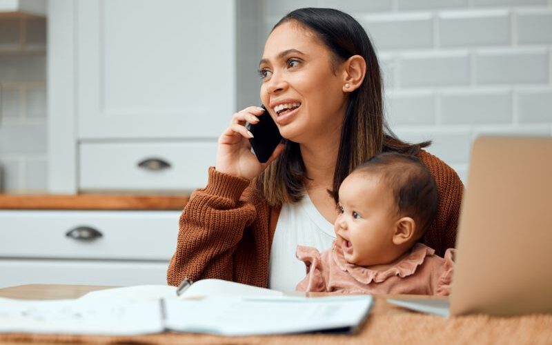 Mother sits at a table holding her baby while talking on a phone.