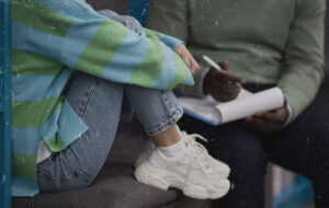 A teen sits on a couch hugging their legs across from a therapist.