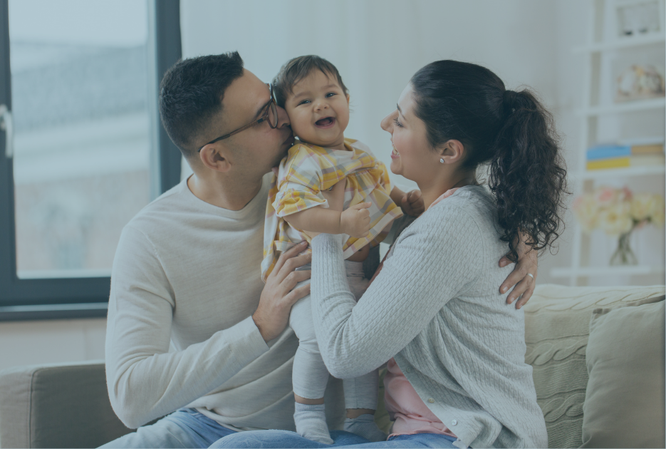 Happy parents sit with baby on couch and embrace her.