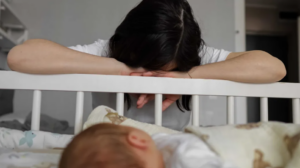 Mother laying her head on a crib next to her sleeping baby.