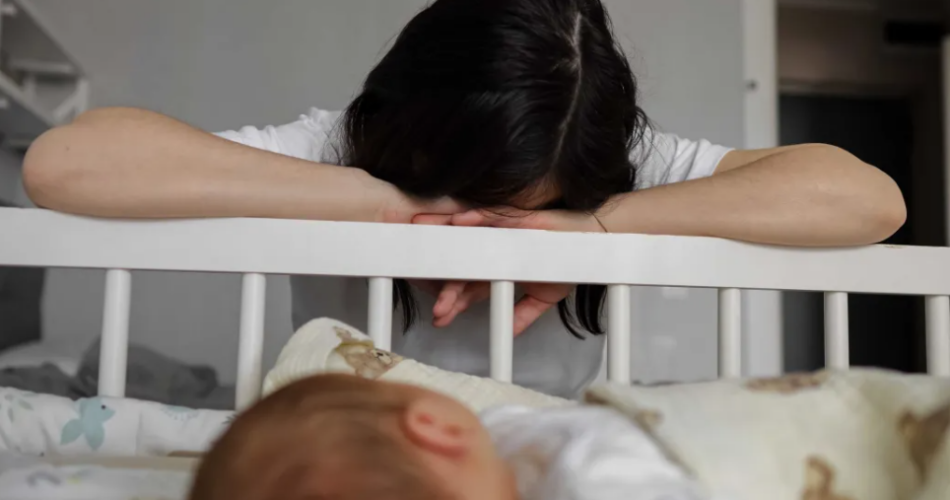 Mother laying her head on a crib next to her sleeping baby.