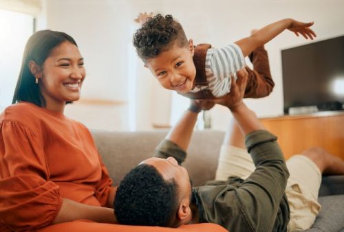 Mother sitting on couch smiling at her husband laying on couch while lifting their son in the air.